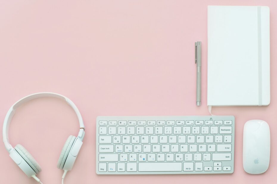 um teclado, um mouse, uma caneta, um bloco de anotações e um headphone em uma superfície rosa